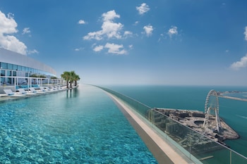 A swimming pool with lounge chairs and umbrellas, surrounded by palm trees and a city skyline in the background.