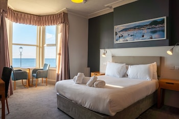 A hotel room with a large bed, two bedside tables, a desk with a chair, a window with curtains, and a painting on the wall.
