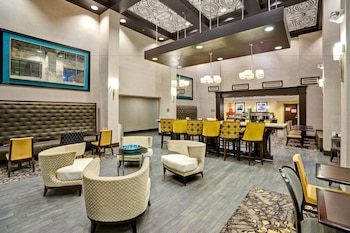 A hotel lobby with a seating area featuring cushioned chairs and tables, a bar area with chairs and a counter, and a large mirror on the wall.