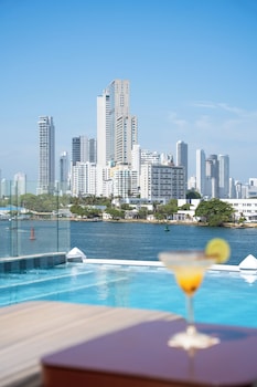 A hotel room with a large bed, a desk, a chair, and a balcony with a view of buildings.