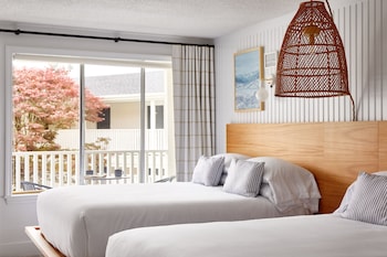 Two beds with crisp linens and striped pillows sit against a wall with vertical paneling, offering a view of a balcony and a serene outdoor setting.