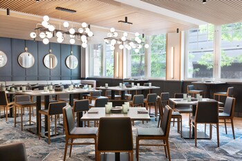 A modern restaurant with round tables and chairs, large windows, and a wooden ceiling.
