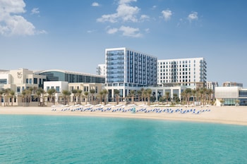 A beachfront hotel with a modern design, featuring multiple balconies, a pool area, and a beach with sun loungers.