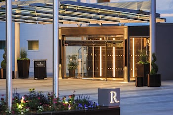Entrada moderna de un hotel con fachada de vidrio, una cubierta metálica y un letrero con la letra 'R'.