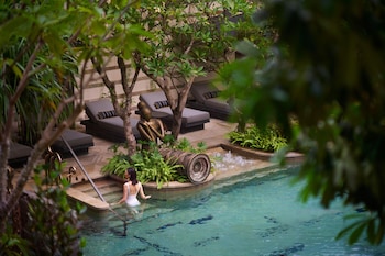 Une personne est assise sur un banc près d’un élément décoratif aquatique, avec un mur de pierre et d’autres éléments décoratifs en arrière-plan.