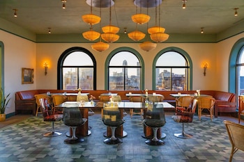 A room with a checkered floor, leather armchairs, a sofa, and a small table with a glass. There are windows with arched tops and a view of buildings outside.