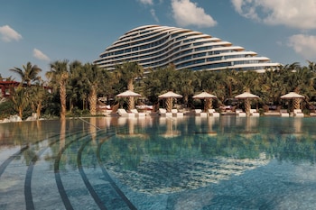 A modern hotel with a unique curved architecture, a pool area with lounge chairs and umbrellas, and palm trees.