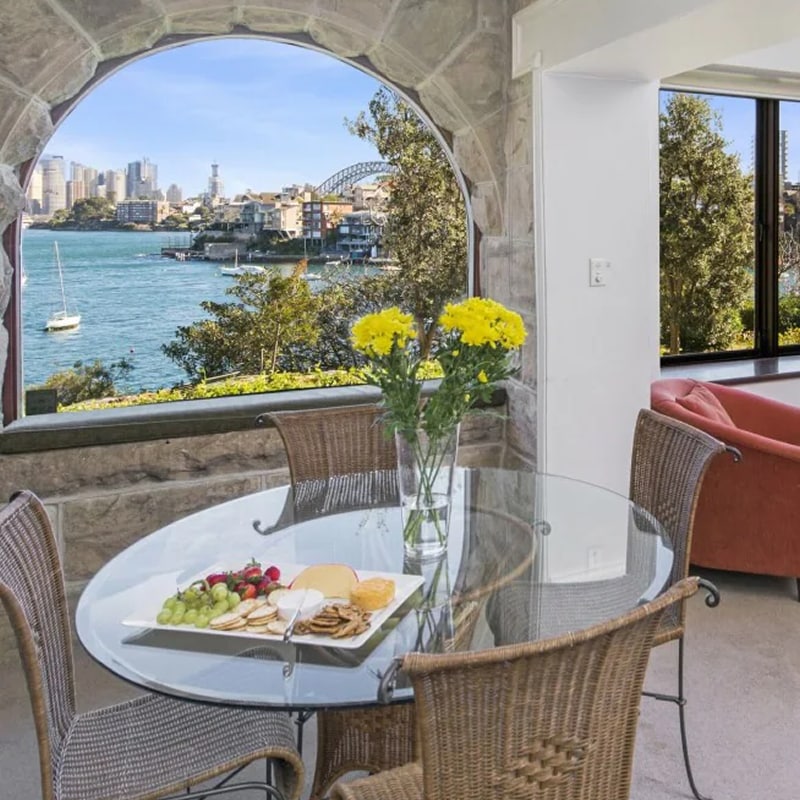 A living and dining space in a Sydney vacation rental, with stone walls in the dining area and a view from the window of Port Jackson and Sydney Harbour Bridge. 