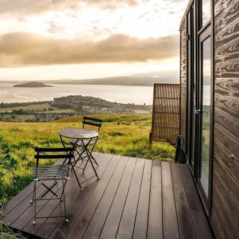 Two dark wood chairs and a table sit on the porch of a wooden cabin with a view of the Bay of Plenty in New Zealand’s North Island. 