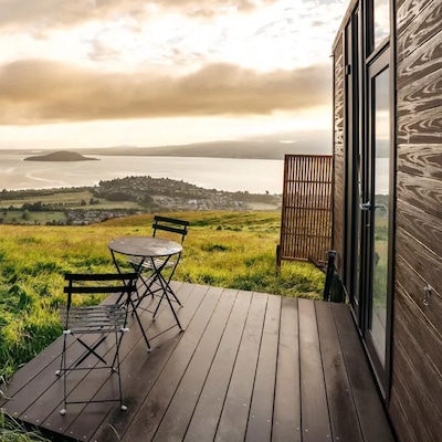 Two dark wood chairs and a table sit on the porch of a wooden cabin with a view of the Bay of Plenty in New Zealand’s North Island.