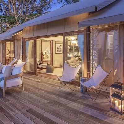 Comfy chairs positioned on a large deck, attached to an inviting holiday home, warmly-lit from inside.