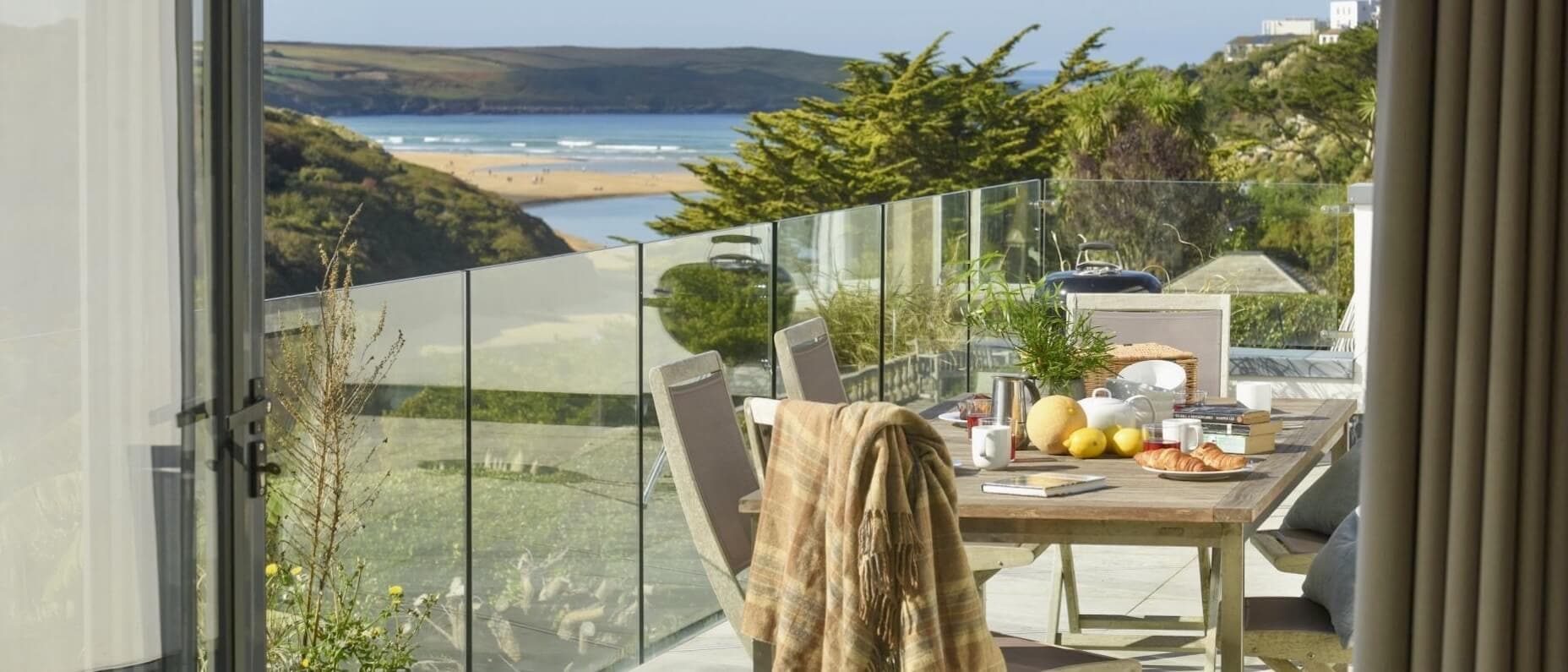 A sunny outdoor dining terrace belonging to a private beachfront home. Breakfast is on the table and there is a view of the sea and hills.