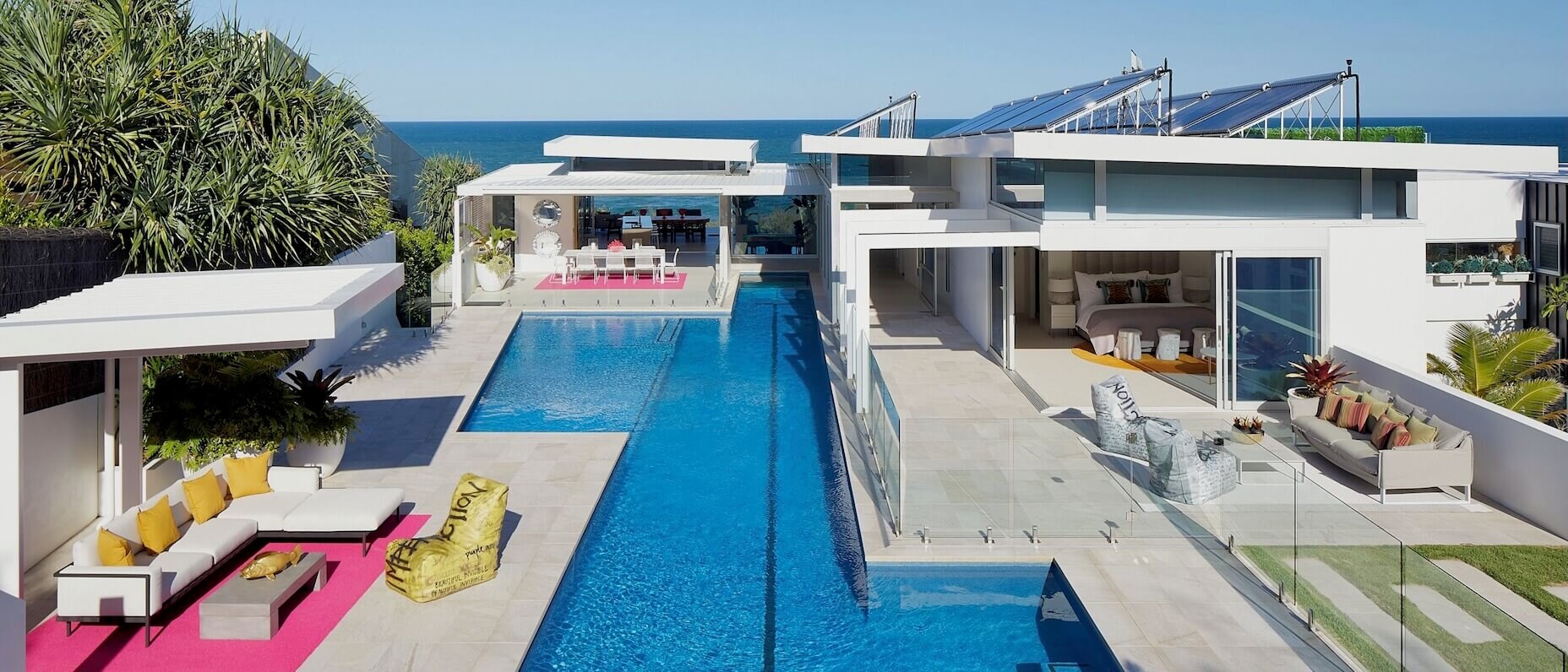 A modern beachfront property with an outdoor pool, multiple seating areas and lush greenery. There is a view of the sea in the background.