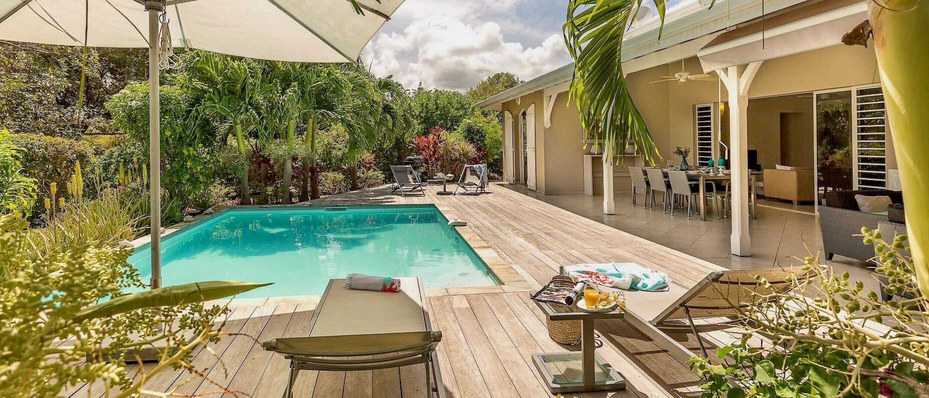 Eine Unterkunft mit Pflanzen rund um einen Außenpool, einem überdachten Sitzbereich und Sonnenliegen. Am blauen Himmel sind ein paar Wolken. 