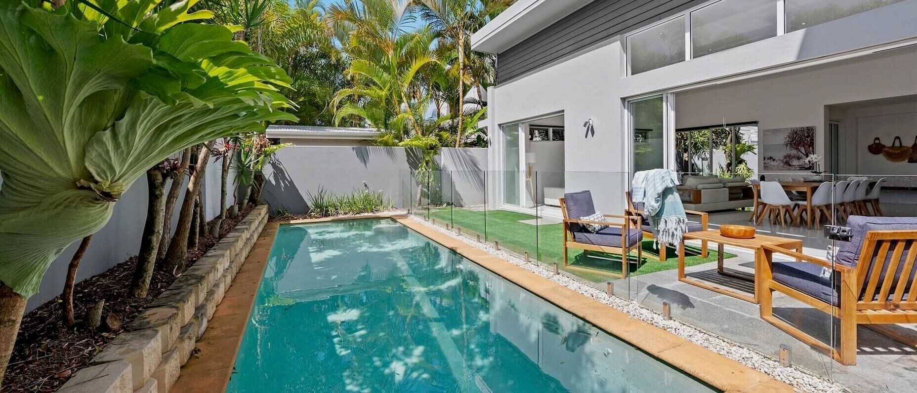 A modern property with an outdoor seating area and a shaded pool surrounded by lush greenery and a blue sky above.