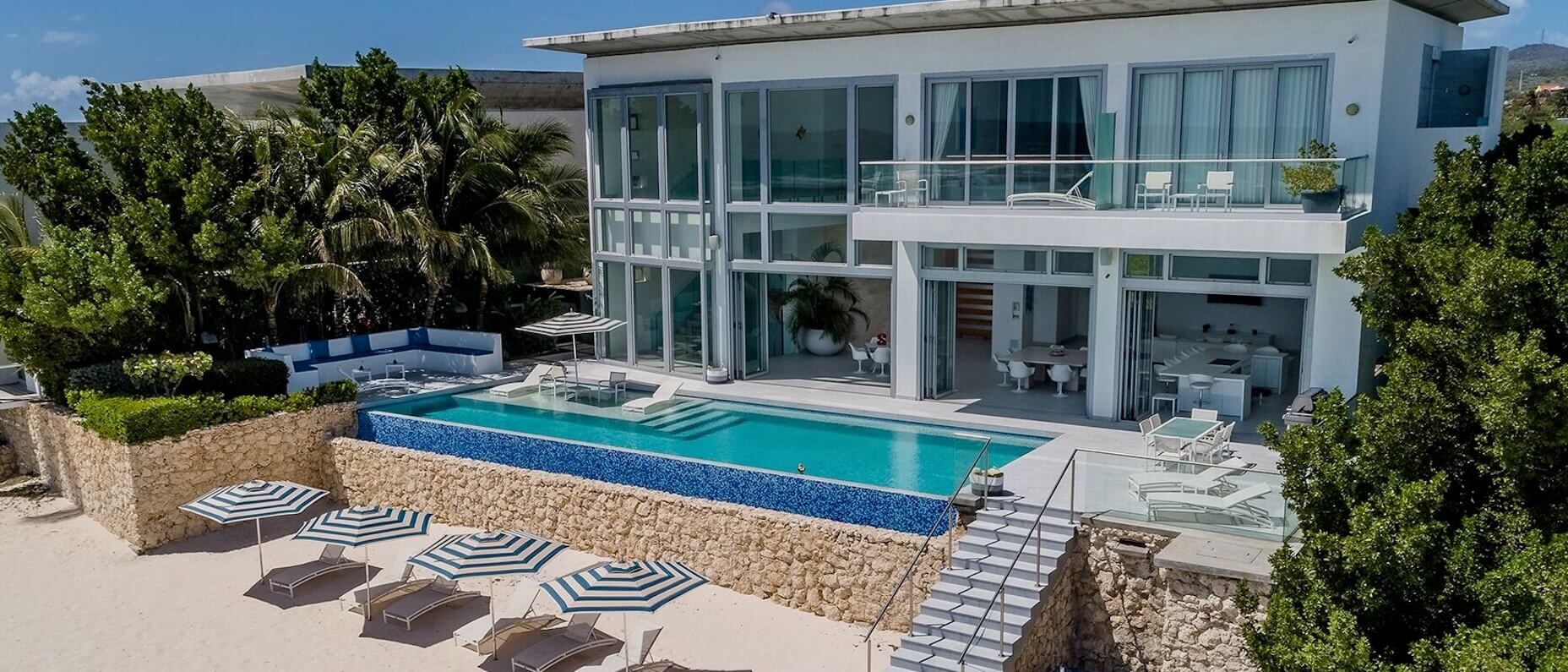 Un hébergement avec de grandes fenêtres, une piscine extérieure, des chaises et des marches menant à une plage ensoleillée.