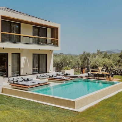 Une maison à deux étages avec piscine privée. Dans le jardin verdoyant se trouvent des lits bain de soleil et un coin repas.