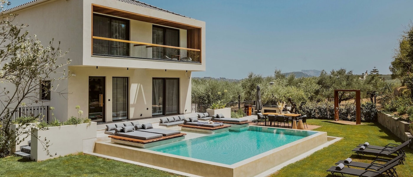 A two-storey home with a private pool in a sunny garden. There are sunloungers, a dining area and greenery surrounding the home.