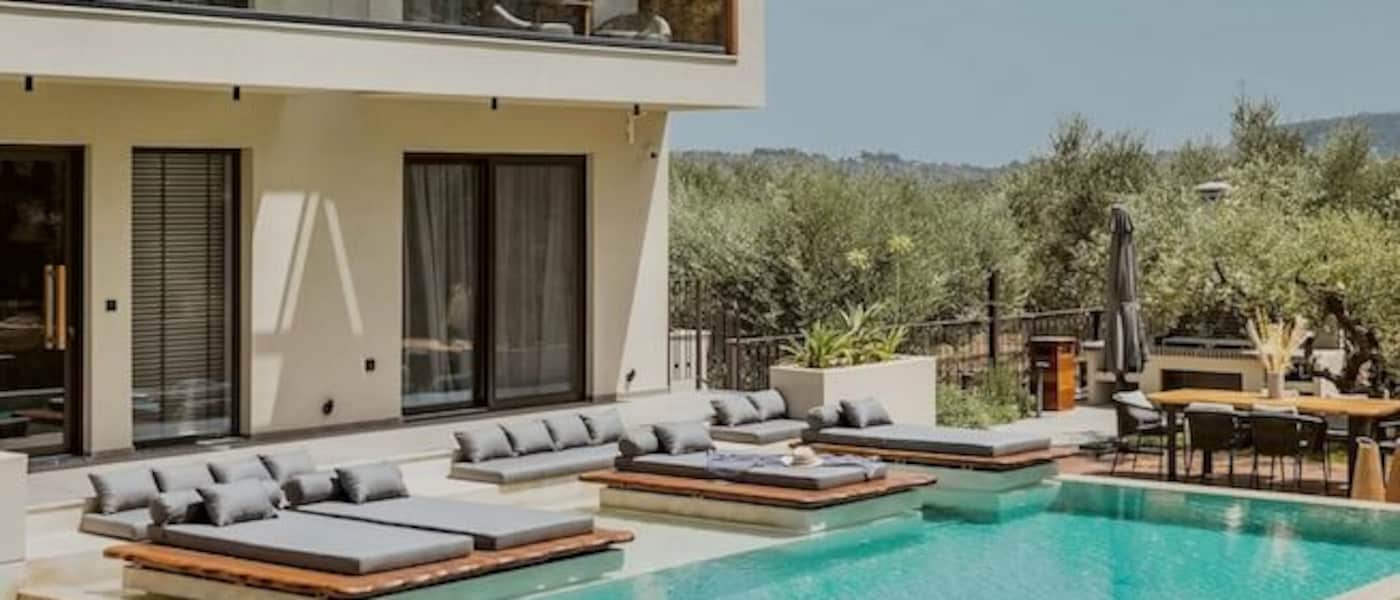 A two-storey home with a private pool in a sunny garden. There are sunloungers, a dining area and greenery surrounding the home.