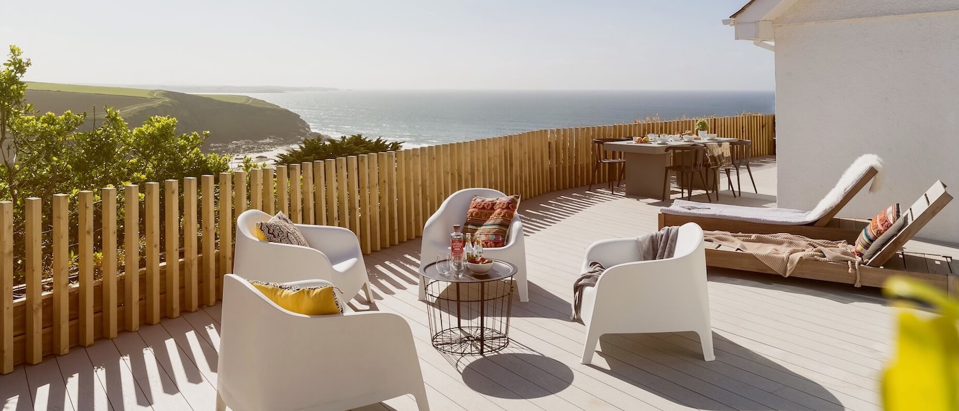 White and brown chairs sit on a pale wood patio overlooking greenery-covered hills and the ocean.