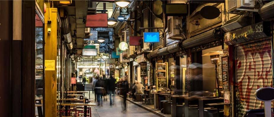 Busy alleyway with shops, cafes, hanging signs, and people at outdoor tables. Walls feature graffiti and colorful decor.