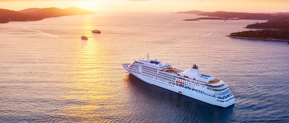 A white cruise ship sailing through calm waters at sunset, with small islands and other boats in the distance.
