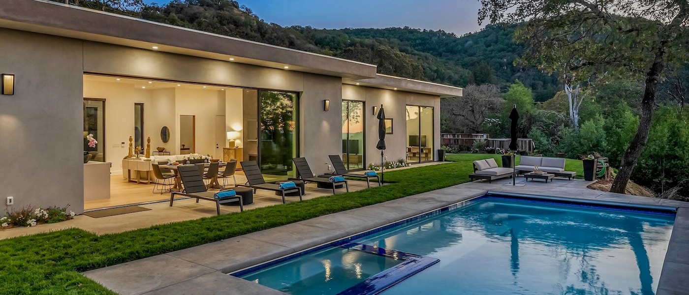 A home with a swimming pool surrounded by green trees and a foliage. Beside the pool, there are a number of seating options.