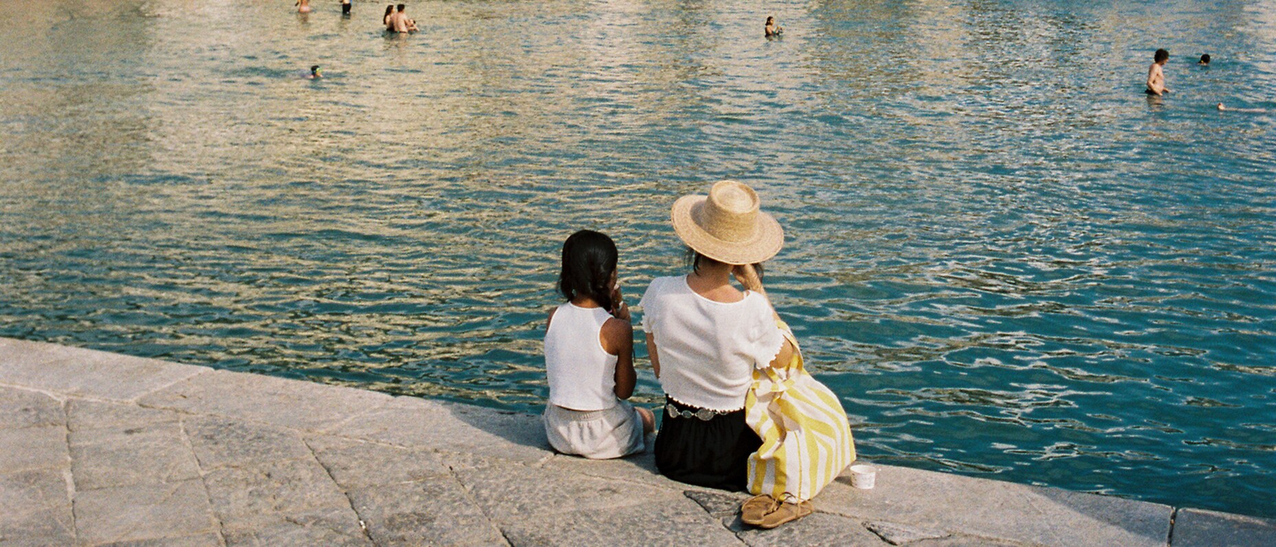 A woman and girl sit on a stone pier, facing a bay with swimmers and old seaside buildings backed by cliffs and hills.A woman and girl sit on a stone pier, facing a bay with swimmers and old seaside buildings backed by cliffs and hills.A woman and girl sit on a stone pier, facing a bay with swimmers and old seaside buildings backed by cliffs and hills.