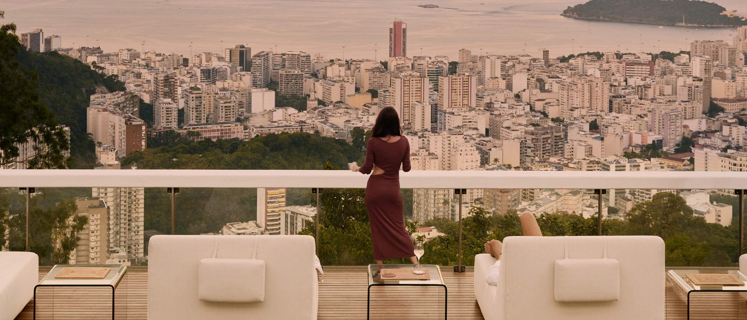 Une personne vêtue d’une longue robe se tient sur une terrasse sur le toit, surplombant un paysage urbain tentaculaire et une baie au coucher du soleil. La terrasse est équipée de chaises longues blanches et d’une piscine au premier plan, avec des montagnes et des îles visibles au loin sous un ciel rose pâle.