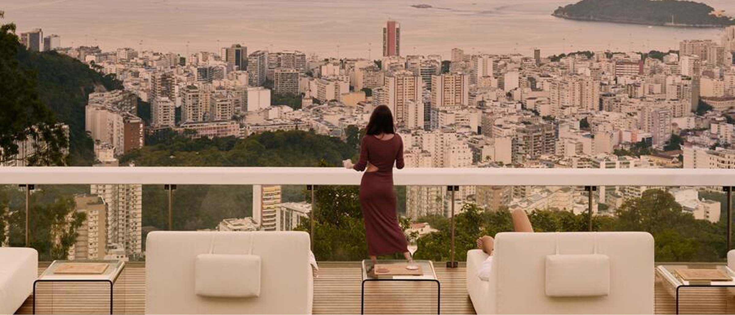 A woman in a long dress stands on a rooftop terrace overlooking a sprawling cityscape and bay at sunset. The terrace features white lounge chairs and a pool in the foreground, with mountains and islands visible in the distance under a soft pink sky.