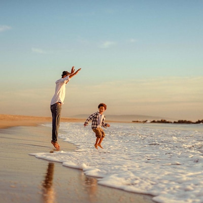 A man and a young boy joyfully play at the edge of the ocean, jumping over waves on a sandy beach during sunset, with the sky glowing in warm tones.