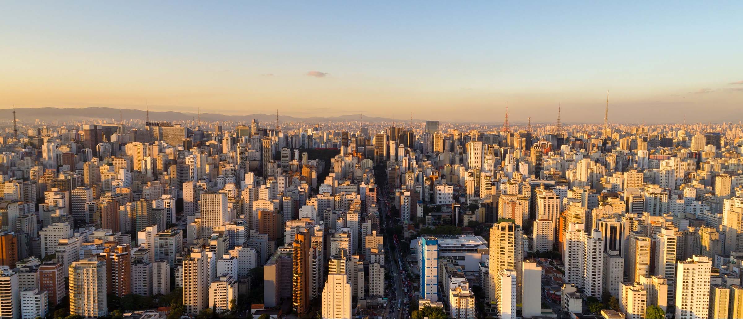 Panoramic view of a modern city skyline with numerous high-rise buildings along the waterfront, framed by lush green trees in the foreground, with the ocean visible in the background.
