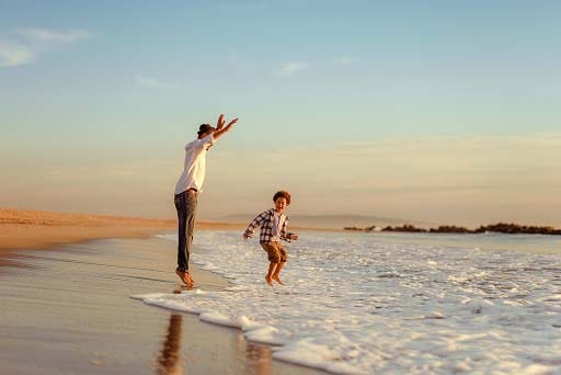 Une personne et un enfant jouent dans l’océan, sur une plage de sable, en sautant au-dessus des vagues au coucher du soleil.