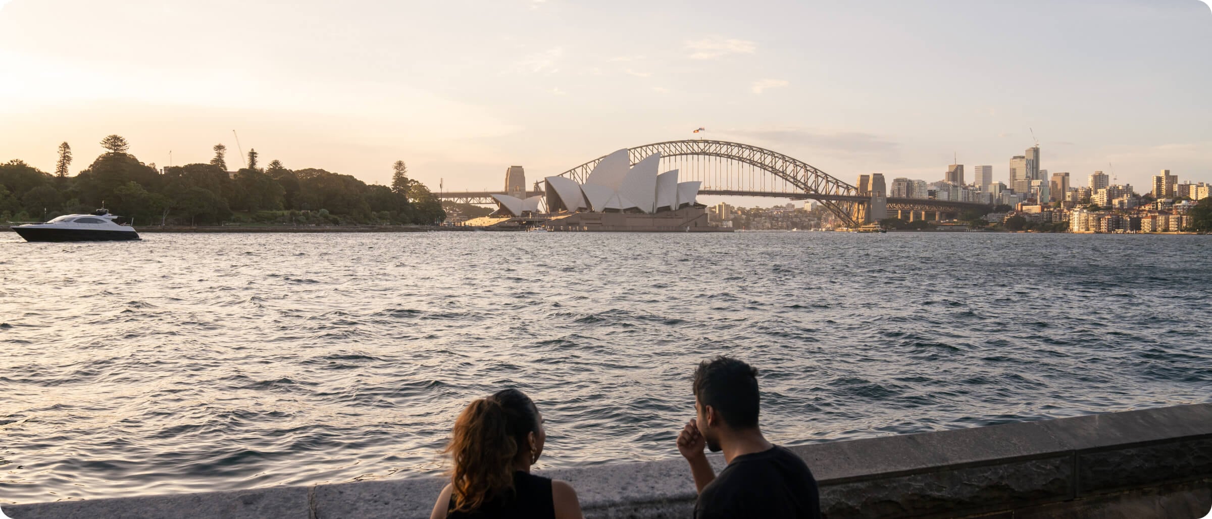 A couple sits by the water at sunset, facing the Sydney Opera House and Harbour Bridge in the distance.