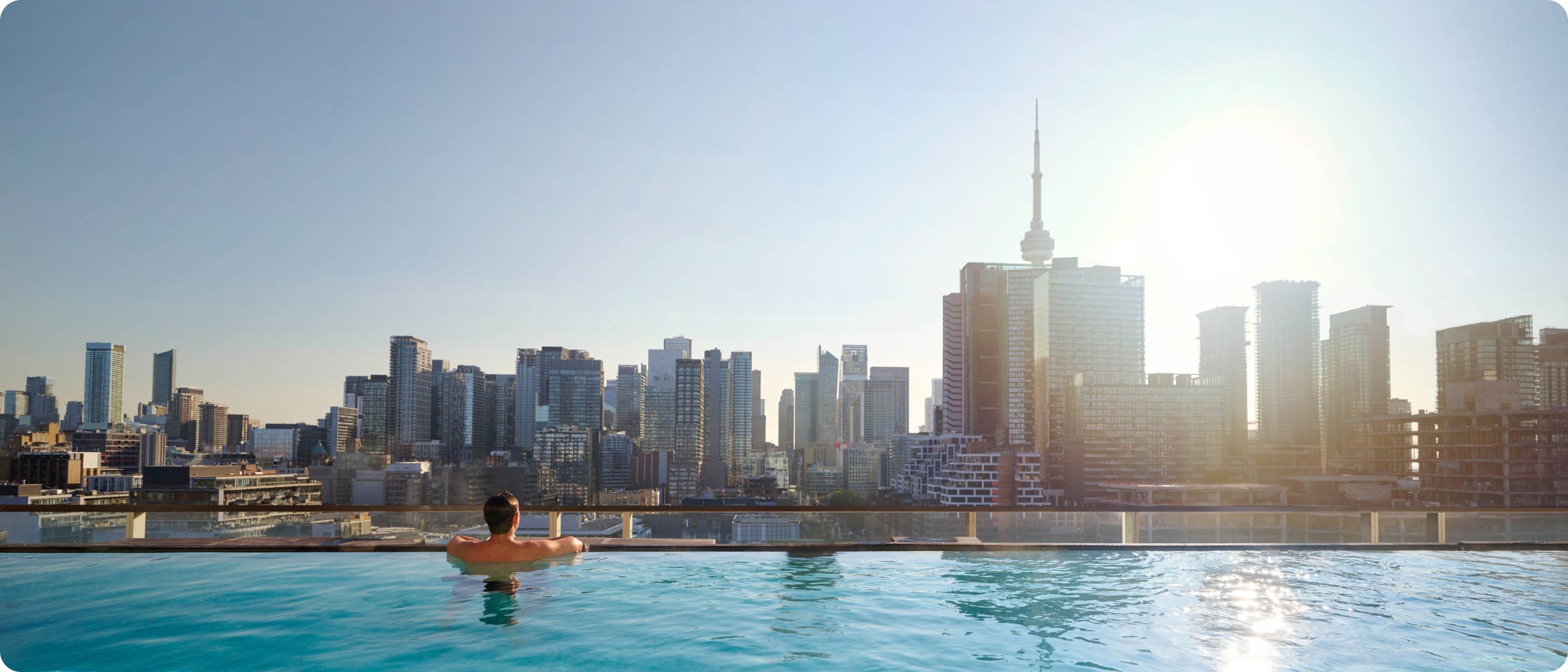 A person enjoys a rooftop infinity pool view with the Toronto skyline and CN Tower visible in the background at sunset.