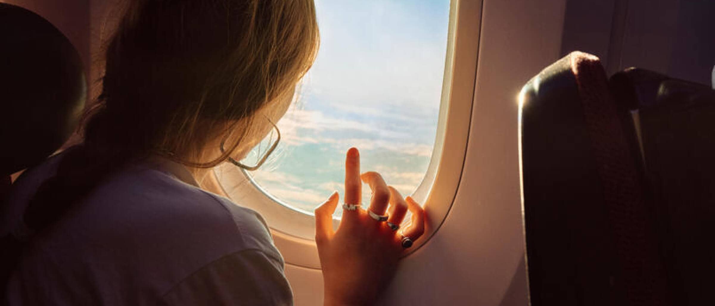 A passenger gazes out of an airplane window, bathed in soft sunlight, with clouds and blue sky visible outside.