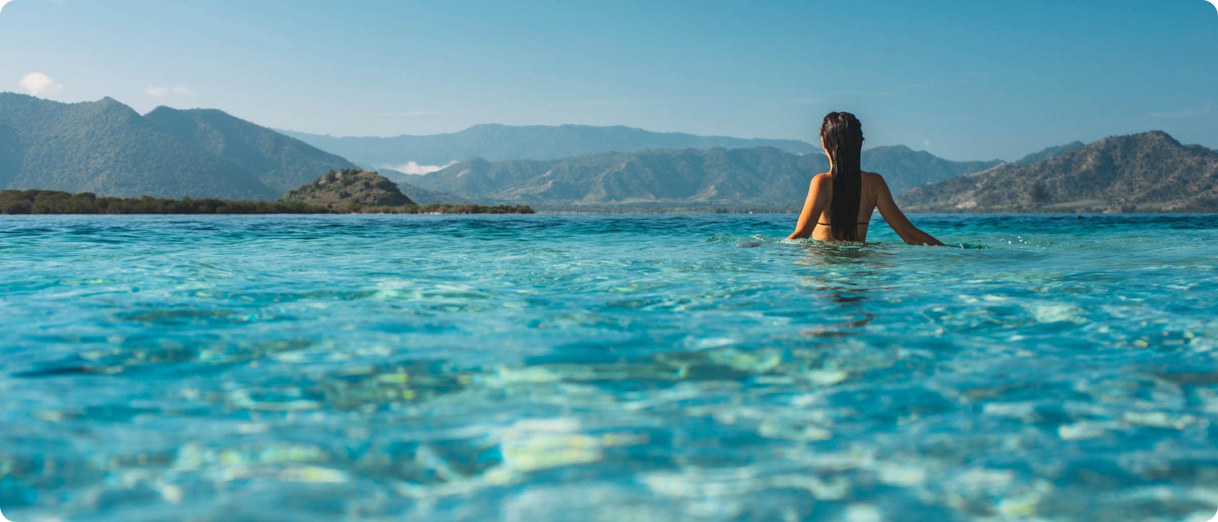 A woman enjoys a serene swim in a crystal-clear infinity pool overlooking mountainous terrain under a blue sky.