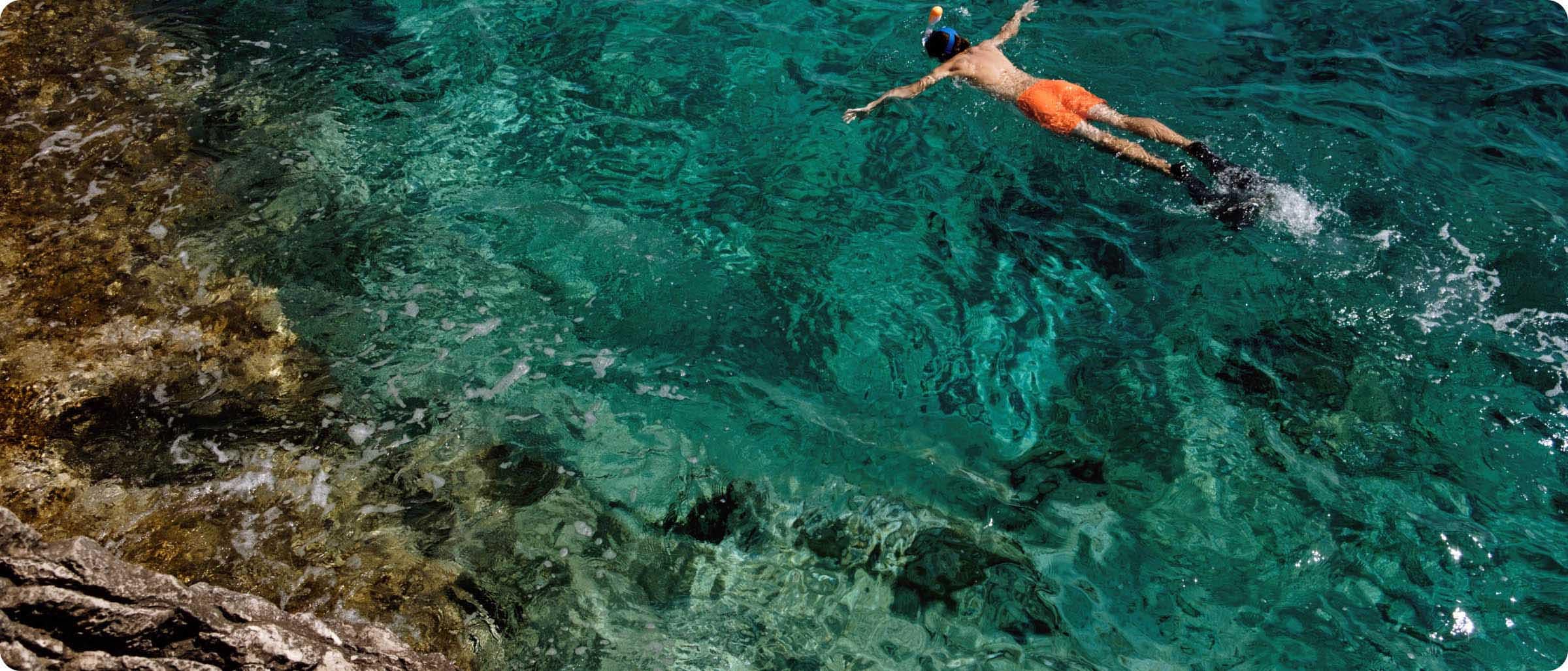 A person snorkels alone in vibrant turquoise waters along a rocky coastline.
