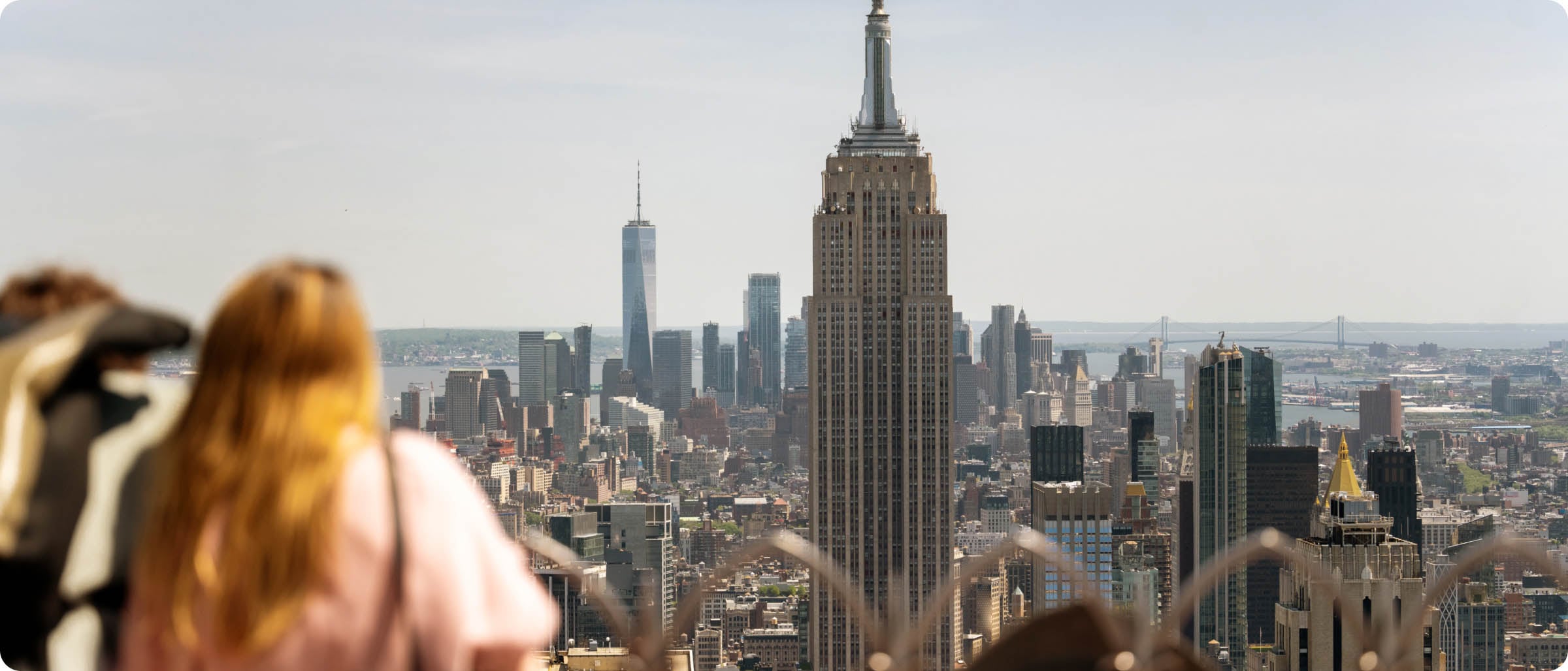Tourists look out over the New York City skyline with the Empire State Building in clear view, surrounded by other skyscrapers under a bright, clear sky.