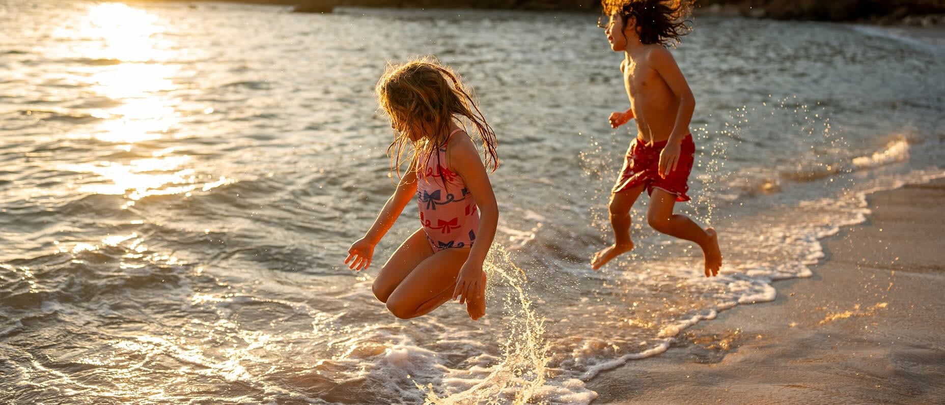 Two children play joyfully at the shoreline, splashing in the shallow waves during golden hour. The sun sets in the background, casting a warm glow over the beach.