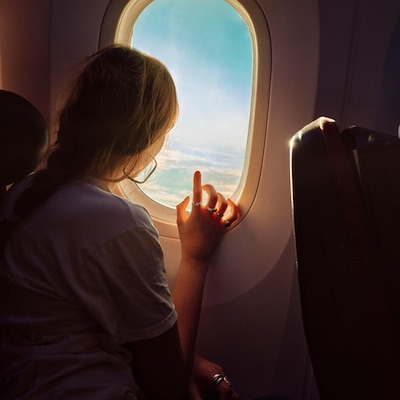 A passenger gazes out of an airplane window, bathed in soft sunlight, with clouds and blue sky visible outside.