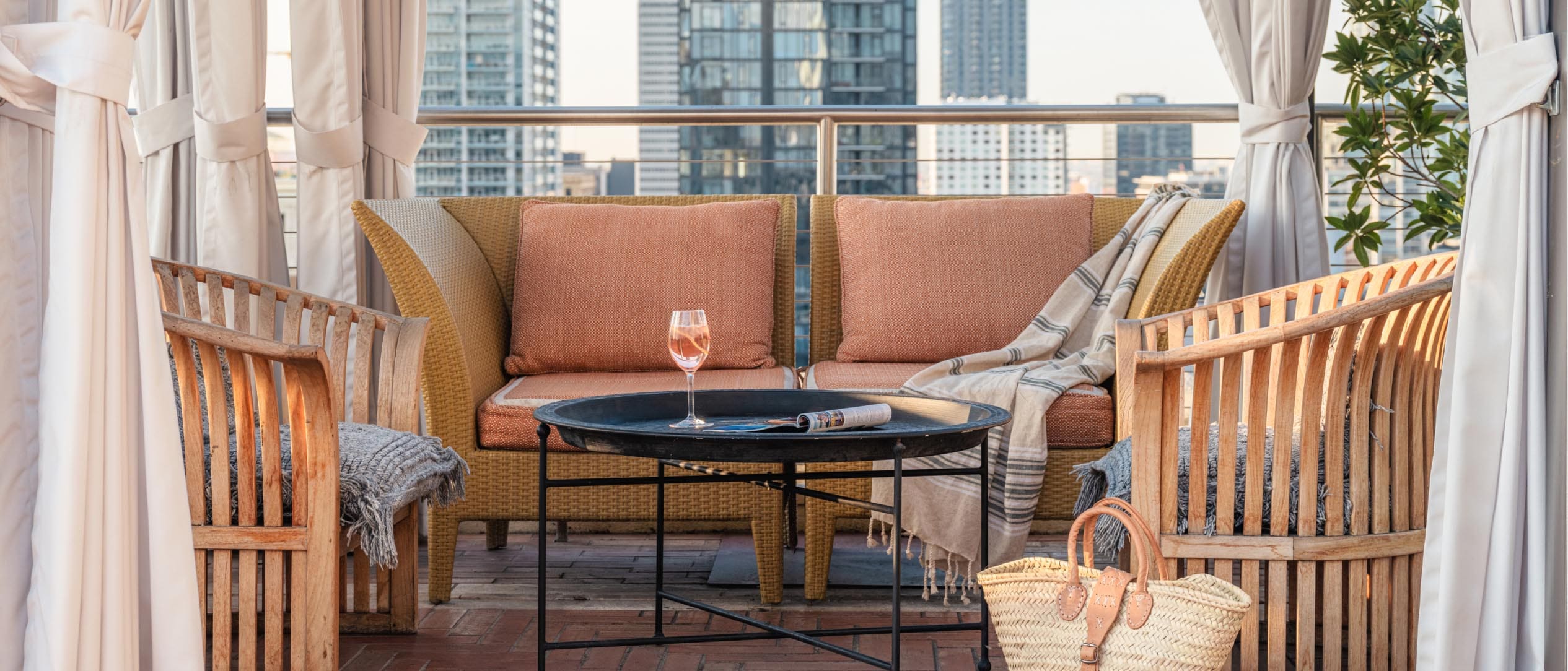 Uma taça de vinho rosé sobre uma mesinha redonda em meio a cadeiras e poltronas ao ar livre com vista para o horizonte da cidade.