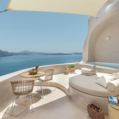 A patio with chairs and an umbrella overlooks an infinity pool facing an ocean and green hills. The sun peeks over the hills.