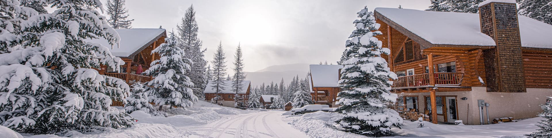 A wooden ski chalet with a snow-covered roof sits before a snowy forest. Behind the chalet, two people ride a chair lift.