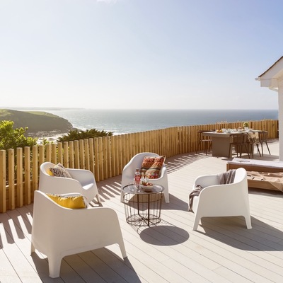 Weiße und braune Stühle stehen auf einem hellen Holzpatio mit Blick auf grüne Hügel und das Meer.