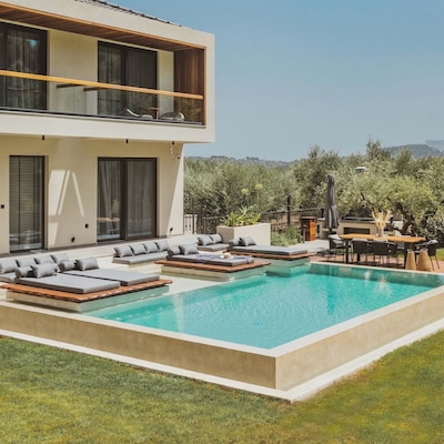 A two-storey home with a private pool in a sunny garden. There are sunloungers, a dining area and greenery surrounding the home.