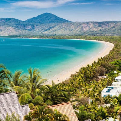 Tropical beach with clear water, palm trees, green mountains, and sailboats. People are along the shoreline.