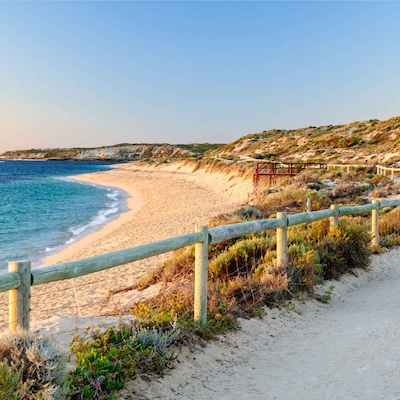 Scenic coastal view with a beach, winding path, grassy dunes, and hills under a clear sky.