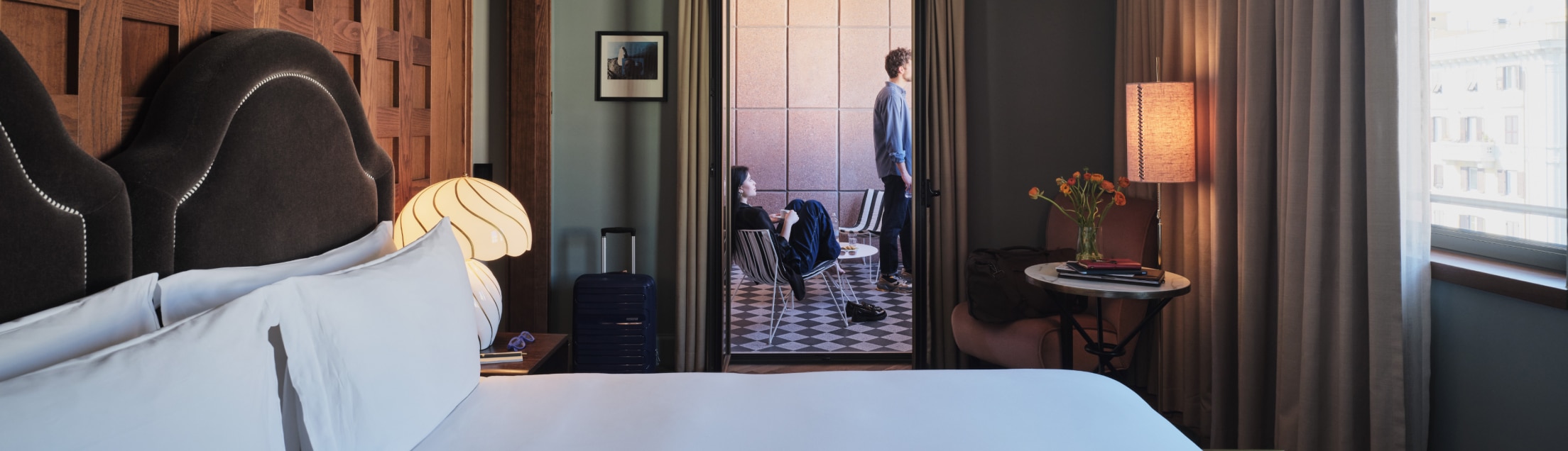 Habitación de hotel con cama en primer plano, mientras un hombre y una mujer se relajan en la sala de estar.