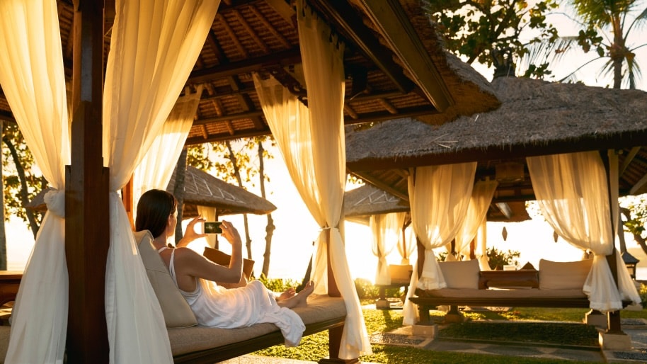 A woman lies in a beach cabana taking a photo of the sunset with her phone.